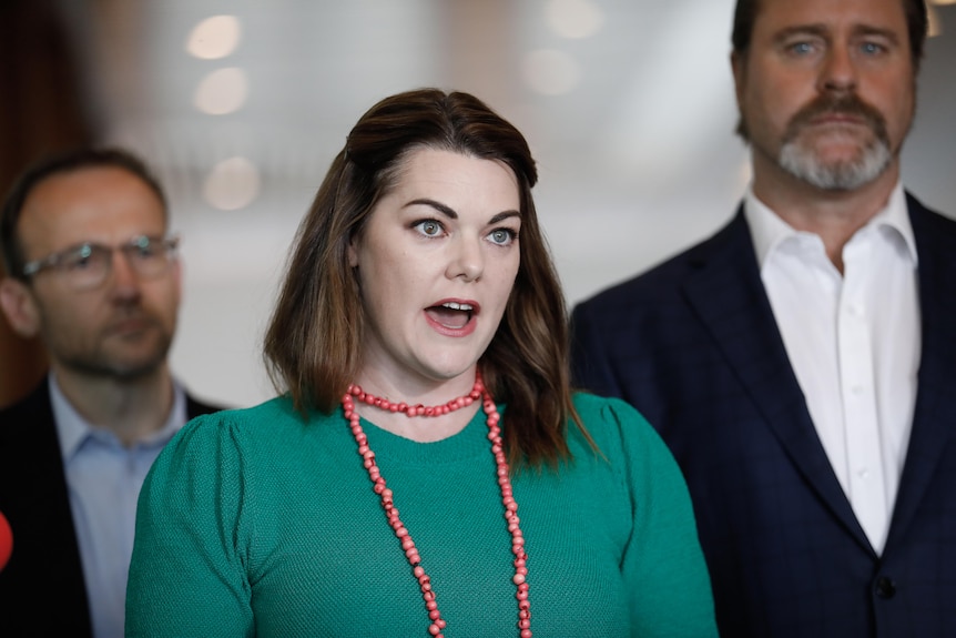 A woman with dark hair – Sarah Hanson-Young – speaks to the media, flanked by men in dark suits.