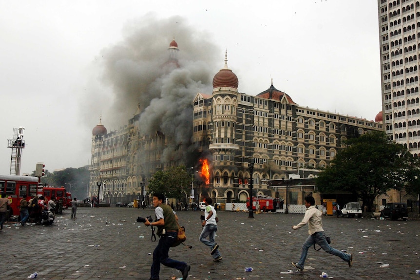 An ornate Indian gothic hotel is shown engulfed by fire as three photographers run across the scene in the foreground.