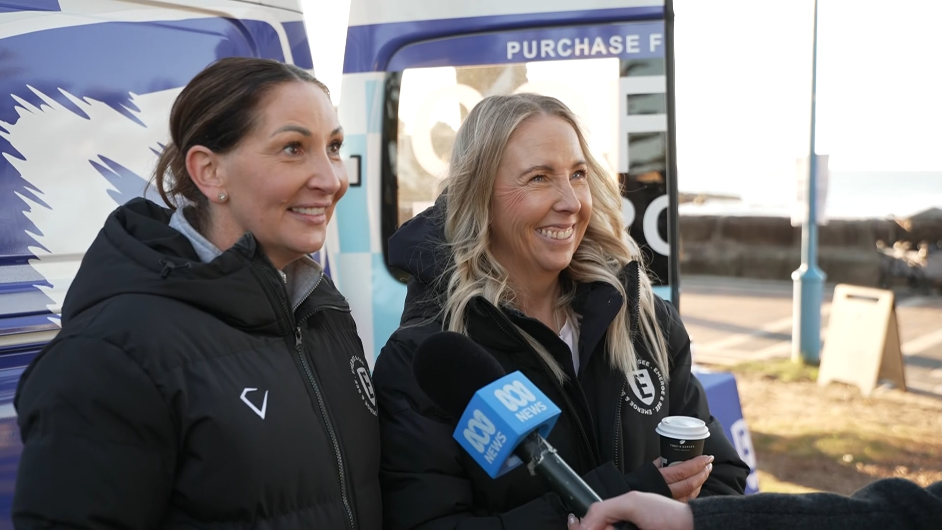Two women smile as one speaks into an ABC microphone.