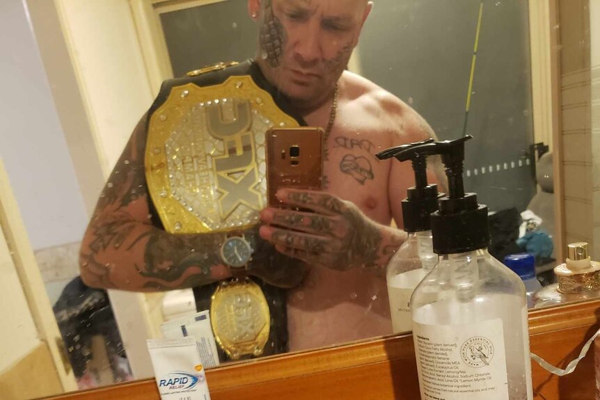 A shirtless, heavily tattooed man with a prize-fighter's belt over his shoulder takes a selfie in a bathroom.