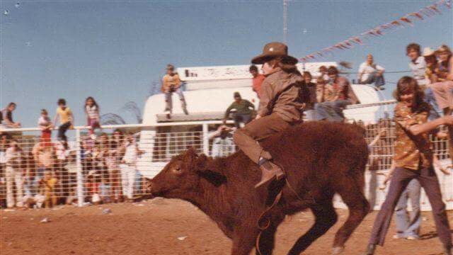 A young boy riding a steer on red dirt in the 1970s.