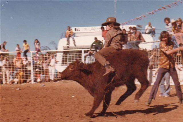 A young boy riding a steer on red dirt in the 1970s.