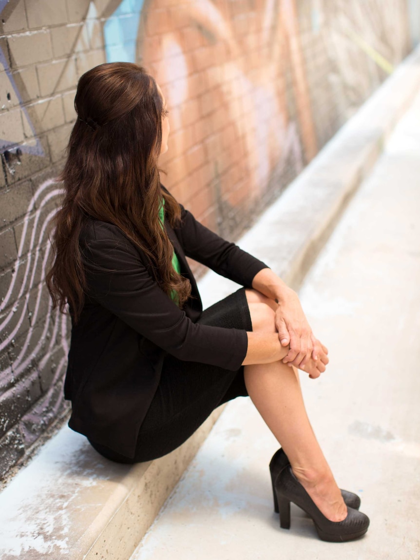 A woman with long brown hair sits on a step, leaving against a brick wall.