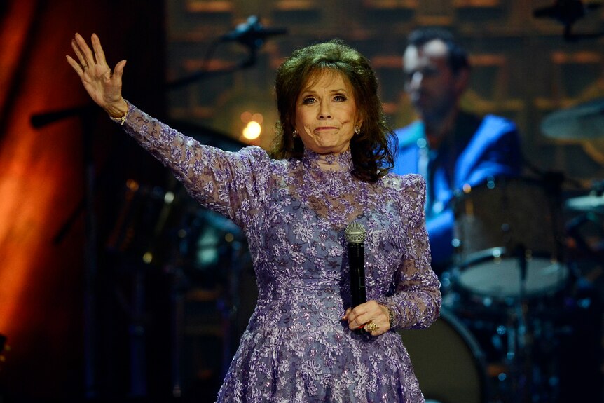 lynn in a purple embroidered gown, mic in hand, waves to the audience