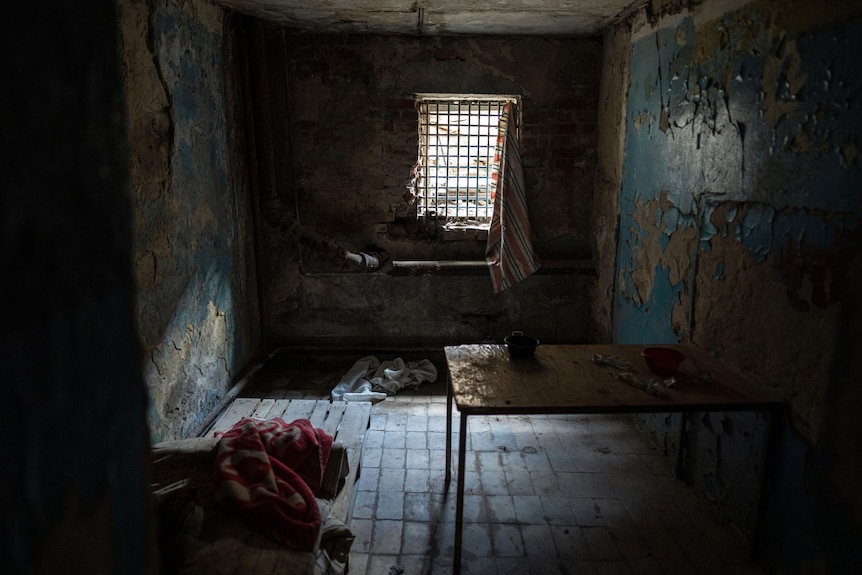 A ramshackle room with bare tiles and peeling paint on the walls, illuminated by light from a barred window.