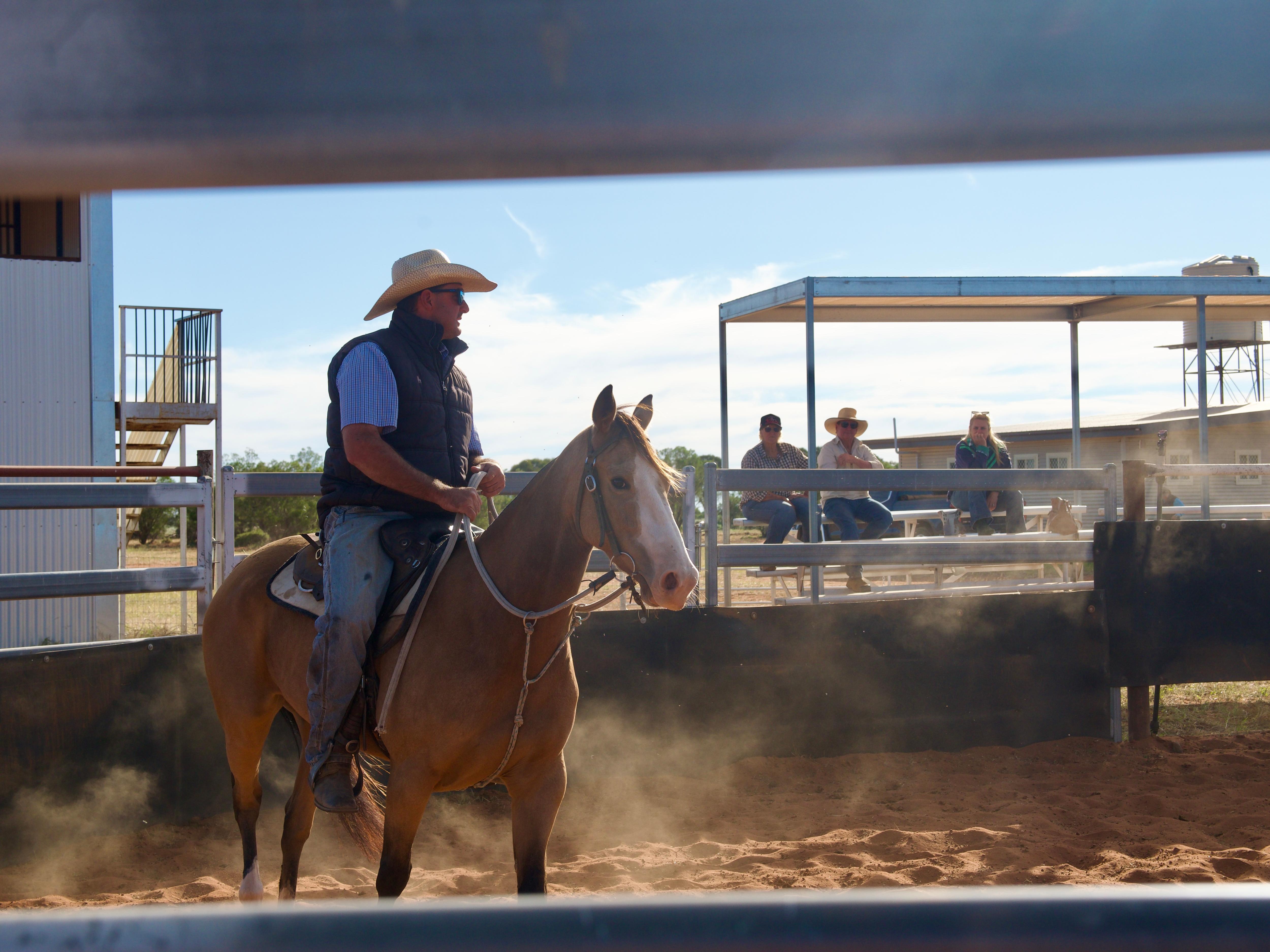 a man with a hat and sunglasses riding a beige horse in a yard with dust 