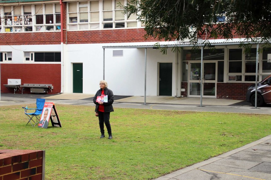 A woman stands in front of a school holding a how to vote card. She is alone.