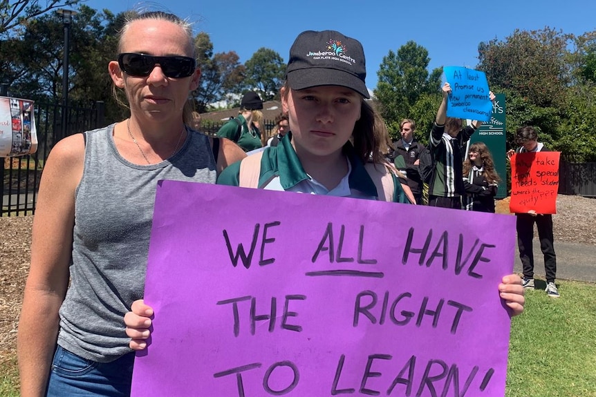 A mother stands beside her daughter holding a sign that reads "we all have a right to learn"