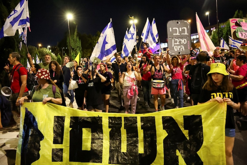 A large crowd of people holding signs in Hebrew. Some are also waving Israeli flags.