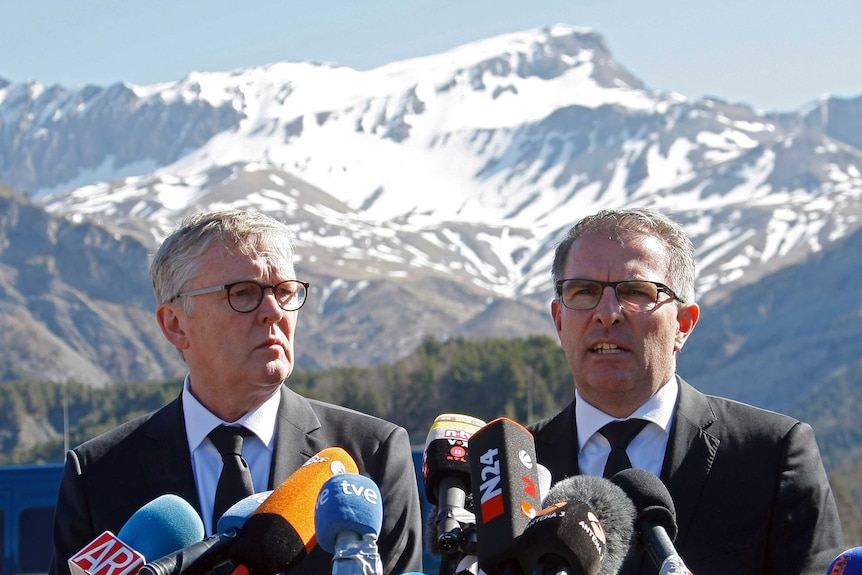 Two men in dark suits and ties, wearing spectacles at a press conference with snow-capped mountains in the background