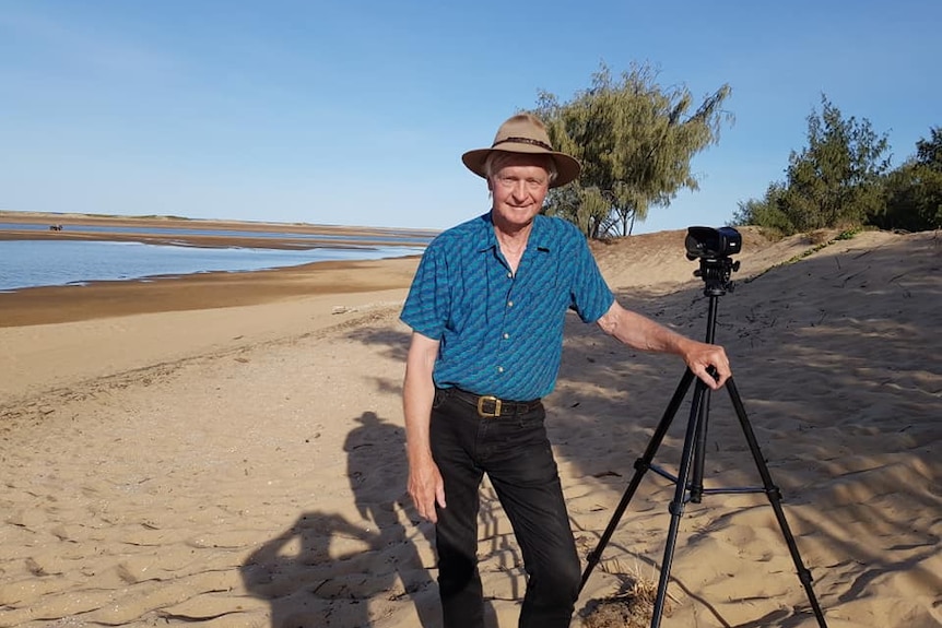 An smiling older man with wide brimmed hat and camera on tripod, at the beach. Wears blue and black stripped shirt, black jeans.