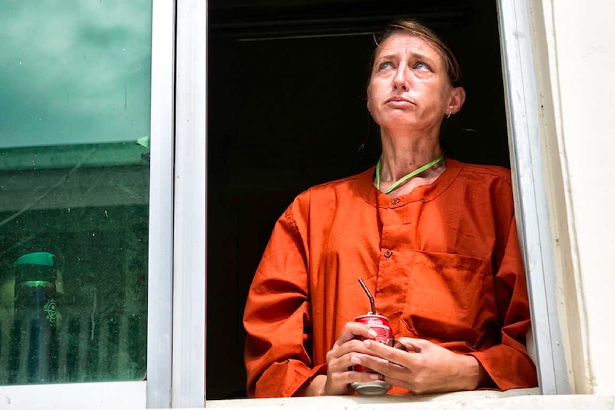 A woman in prison orange uniform looks solemn standing by a window