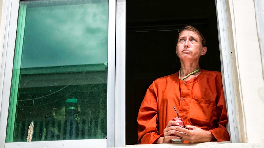 A woman in prison orange uniform looks solemn standing by a window