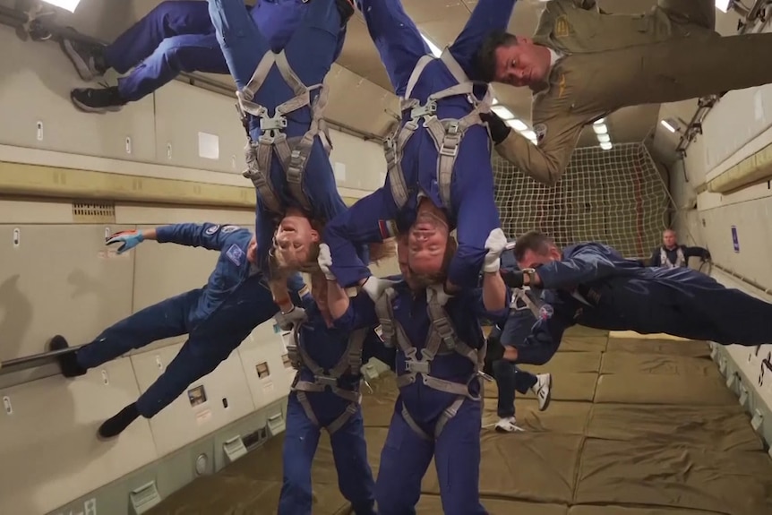 Four people in blue jumpsuits cling to one another as they float in zero-gravity.