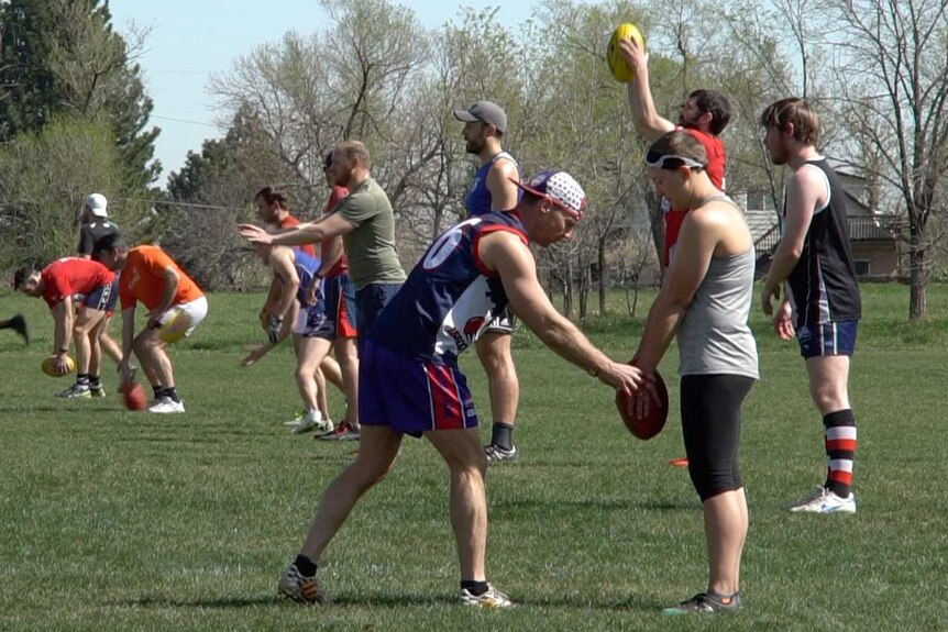 A male Denver Bulldogs player instructs a female player on kicking a football as other male players train in the background.