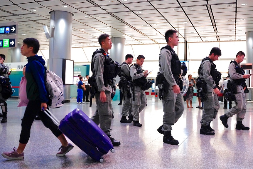 Inside an airport terminal, you view a group of male riot police officers patrolling as one passenger wheels luggage.