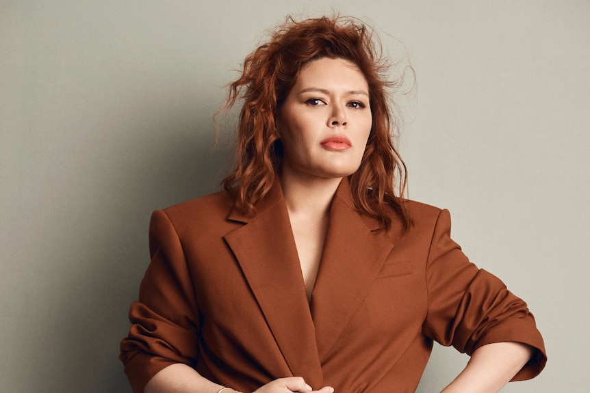 Nakkiah Lui, an Indigenous woman with auburn hair, is wearing a brown blazer dress, looking at the camera.