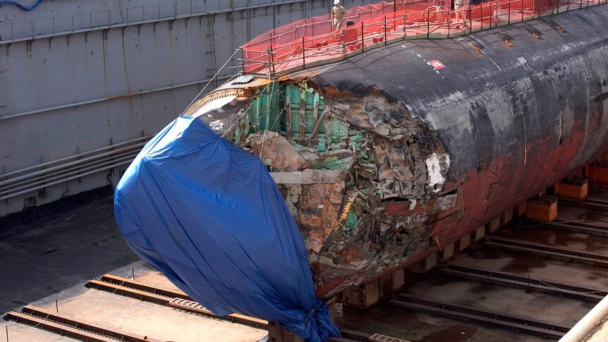 You see a US submarine with its front badly damaged and partially covered by a blue tarp in a dry dock.
