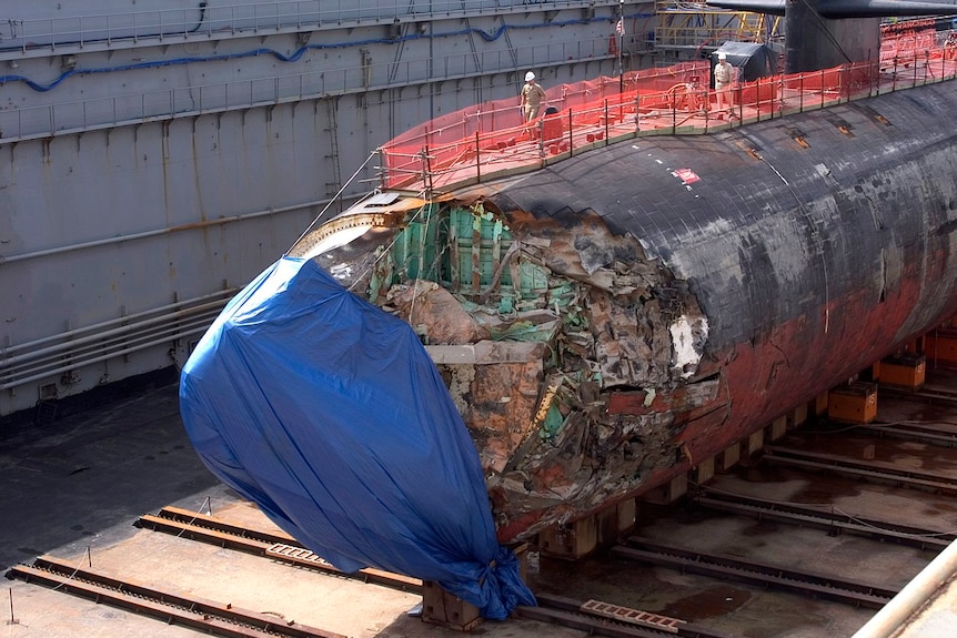 You see a US submarine with its front badly damaged and partially covered by a blue tarp in a dry dock.