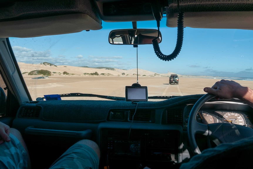 Interior of a four wheel drive vehicle showing driver's hand, passenger legs and big wide beach out the windscreen.