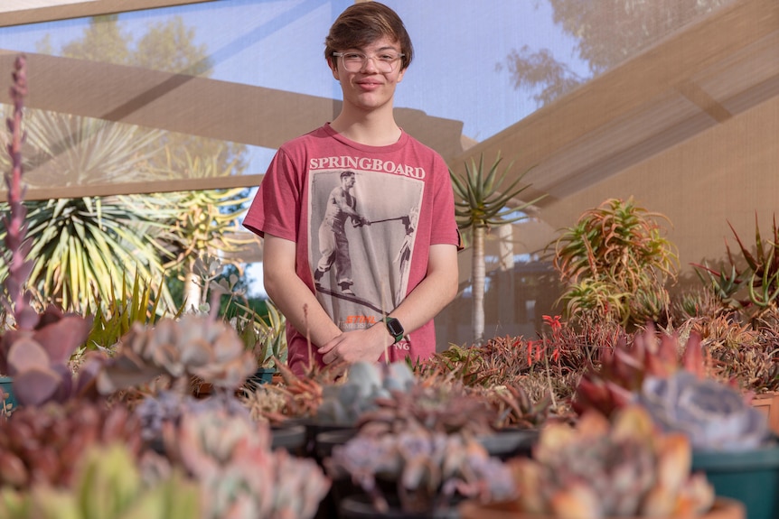 A young white male with brown hair smiling behind a cactus garden, he looks happy