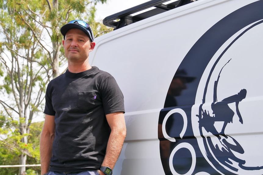 A man wearing a black t shirt, cap and sunglasses standing in front of a white van. 