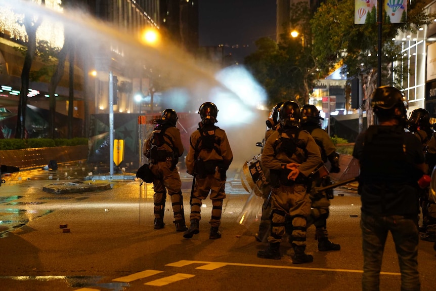Police watch as a water cannon fires on protesters down a street.