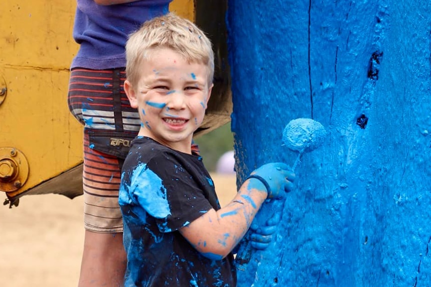 Little boy covered in blue paint painting a tree
