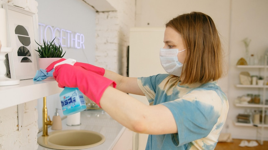 Woman wearing mask and pink gloves cleans her home, in a story about how to clean mould after rain.