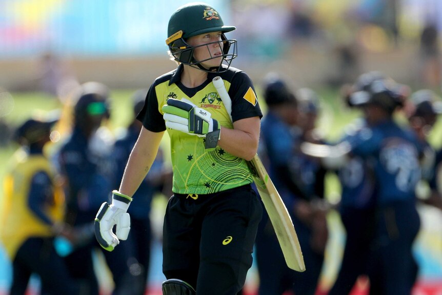 Alyssa Healy holds her bat under her arm and looks off to one side as players celebrate behind her