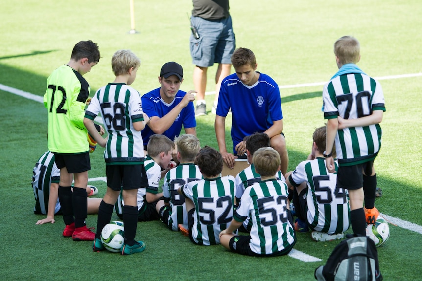 a group of young male soccer players sitting on the football pitch listening to their trainer