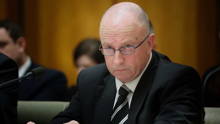 Deputy Commissioner Gaughan sits behind the table, wearing glasses and looking straight ahead.