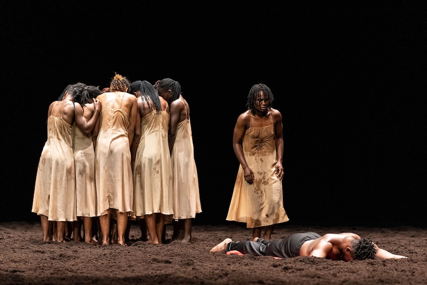 A group of African dancers stand huddled on stage, with one man face-down in the dirt which covers the floor