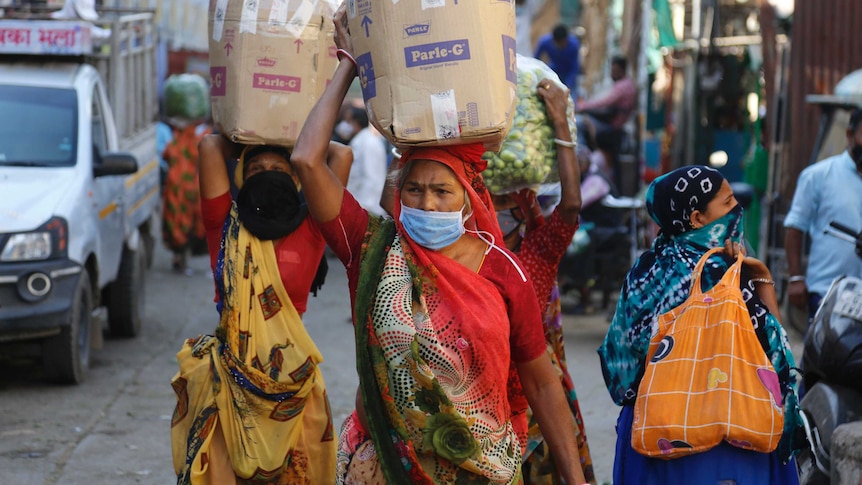 Several brightly dressed women carrying boxes and other goods on their head and wearing surgical masks walk down a street.