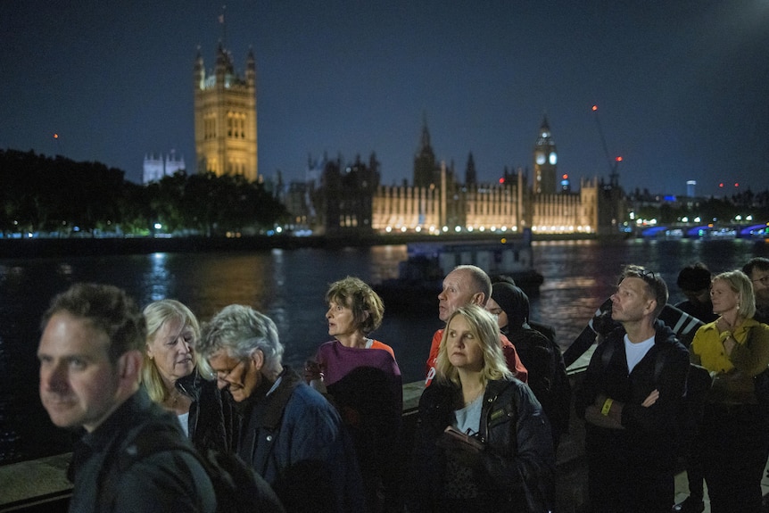 People queue along the Thames in London at night. Westminster is in the background.