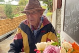 An elderly man wearing a hat sits next to a bouquet of flowers.
