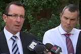 Mark McGowan speaks at a press conference surrounded by microphones as a man in a shirt and tie watches on.