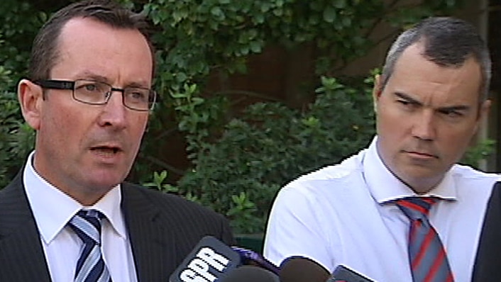Mark McGowan speaks at a press conference surrounded by microphones as a man in a shirt and tie watches on.