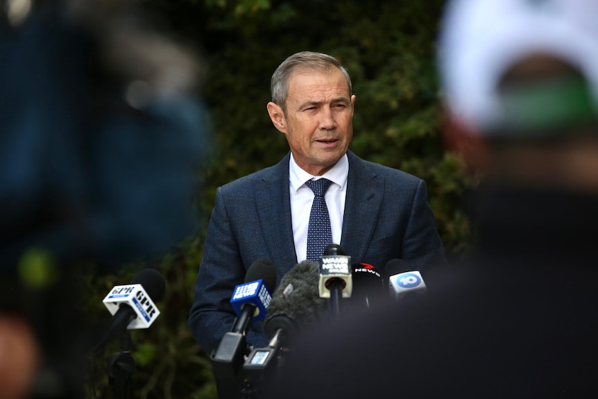 Roger Cook addresses media as he is pictured through the middle of two media people