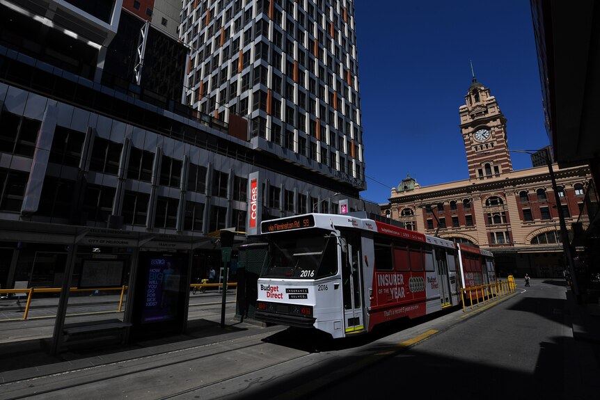 A tram drives in front of Flinders Street station, a large sandstone building, in Melbourne CBD.