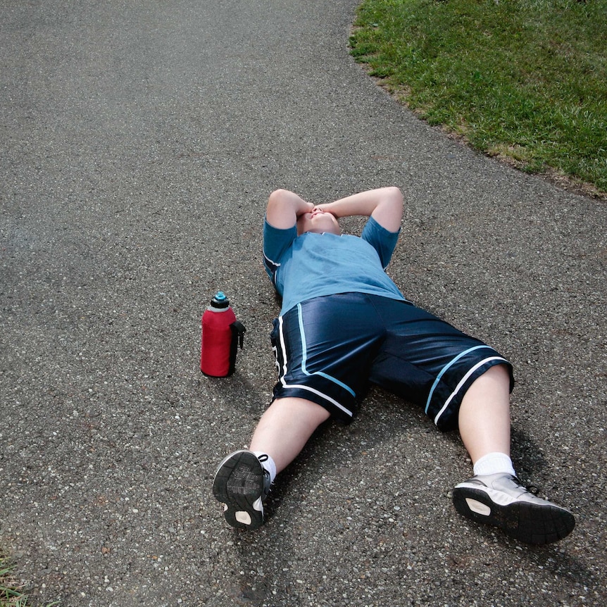 Young boy wearing blue sports top and shorts lying on the road with his hands over his face. There is a red water bottle nearby.