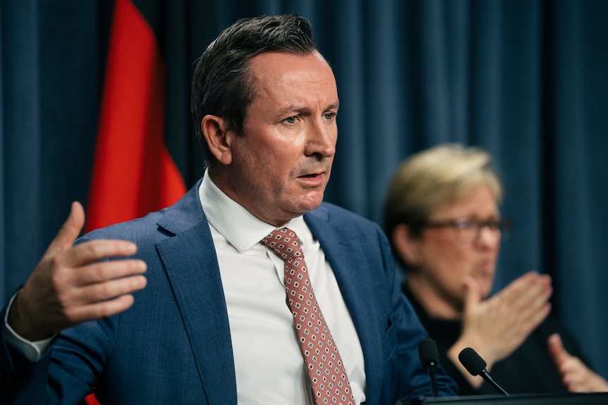 Mark McGowan wears a suit in a room with a blue curtain behind him and australian flags, with a sign language interpreter