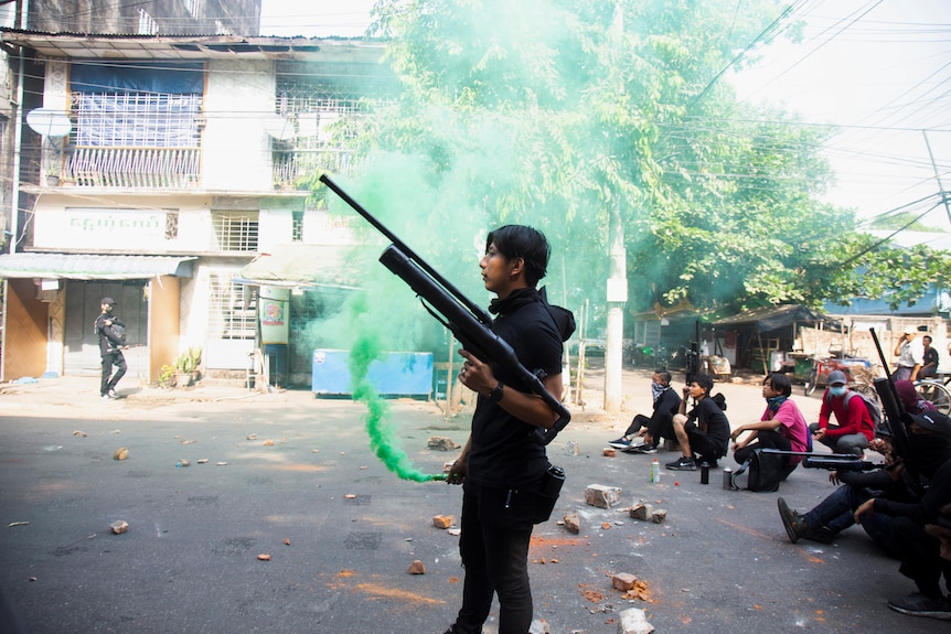 A young Asian man holds a long rifle as people sit behind him in tropical village.