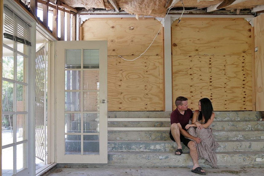 Townsville residents Sarah Little and Chris Baker sit on the stairs of their partially repaired home, a year after it flooded.