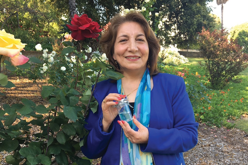 Mahin Nowbahkt, wearing a jacket, standing in a garden next to a rose bush, holding a small jar of rose water.