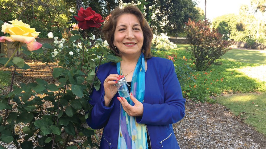 Mahin Nowbahkt, wearing a jacket, standing in a garden next to a rose bush, holding a small jar of rose water.