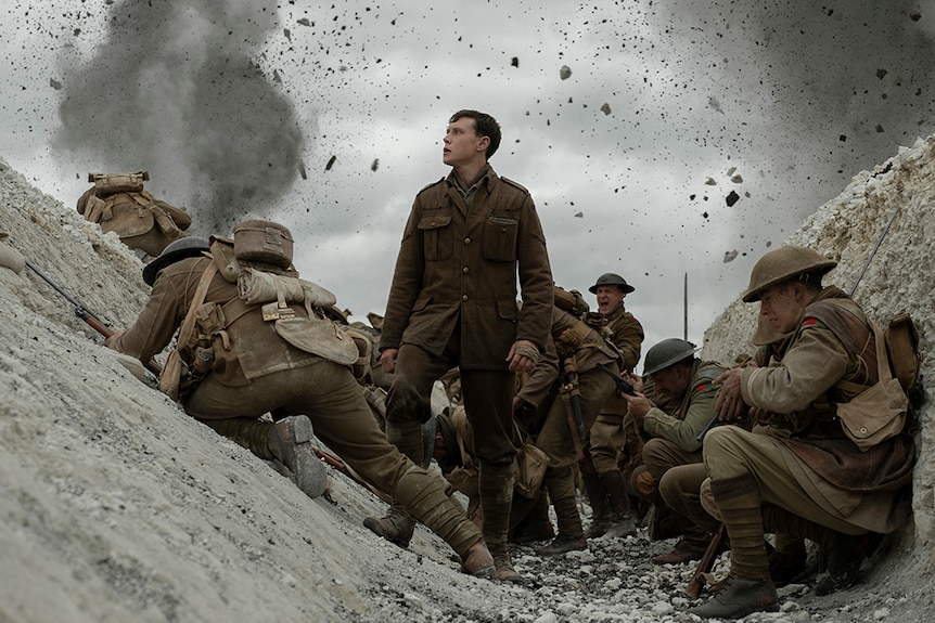 A man in world war one brown uniform stands looking into the distance in the middle of trench full of soldiers under attack.