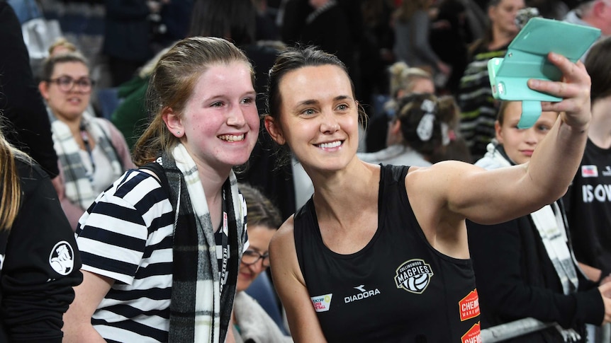 A netballer holds her phone out to take a selfie of her and a beaming young fan at courtside.