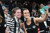 A netballer holds her phone out to take a selfie of her and a beaming young fan at courtside.