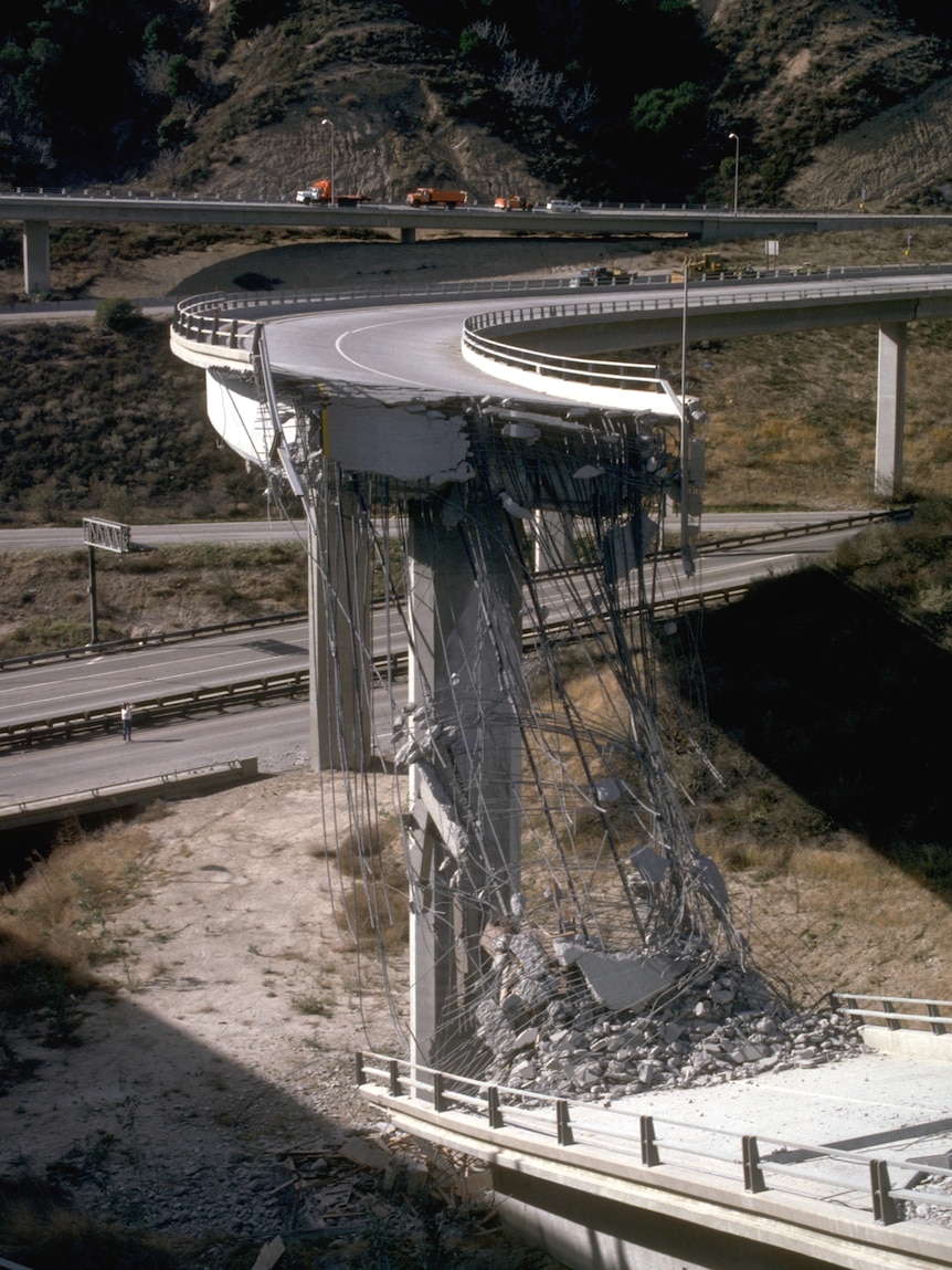 A curved motorway on-ramp is partially destroyed in a hilly landscape with its steel braces hanging off of the remaining ramp.